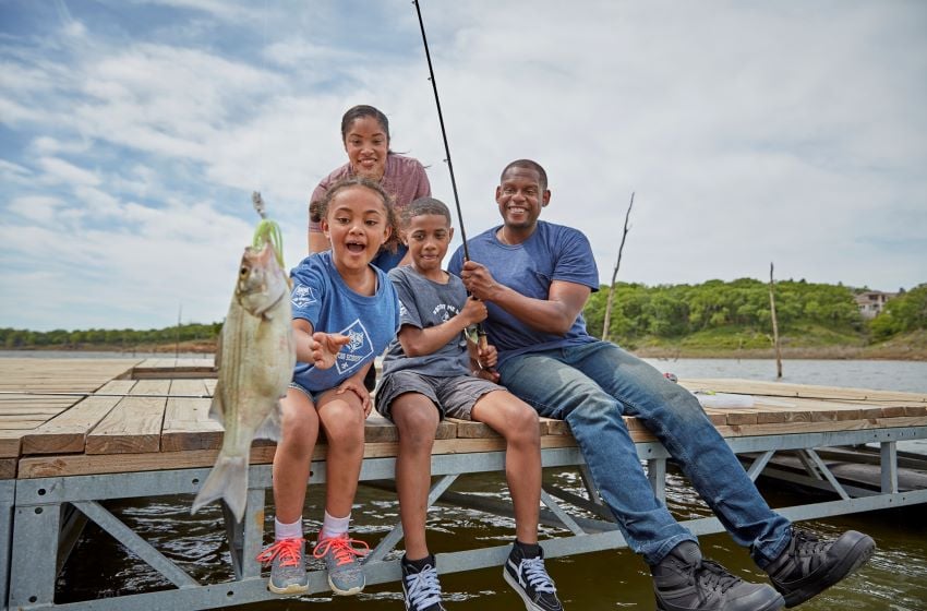 Family sitting on a dock fishing together, smiling as their child proudly holds up a freshly caught fish during a Cub Scout outdoor adventure.