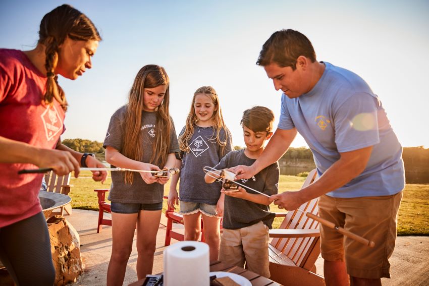 Scouting family roasting marshmallows at sunset around a campfire, laughing and bonding during an outdoor Cub Scout activity.