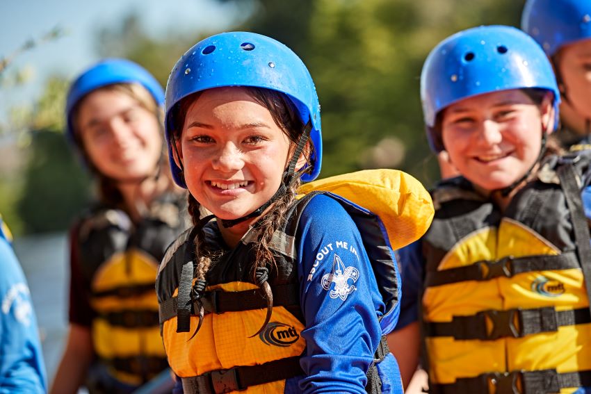 Scouts wearing blue helmets and yellow life vests, smiling during water-based outdoor activities promoting safety and teamwork.