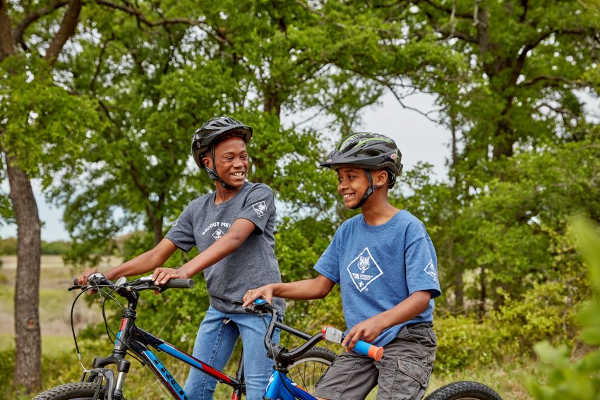 Two Scouts wearing helmets biking through a wooded trail, enjoying outdoor adventure and learning new skills.