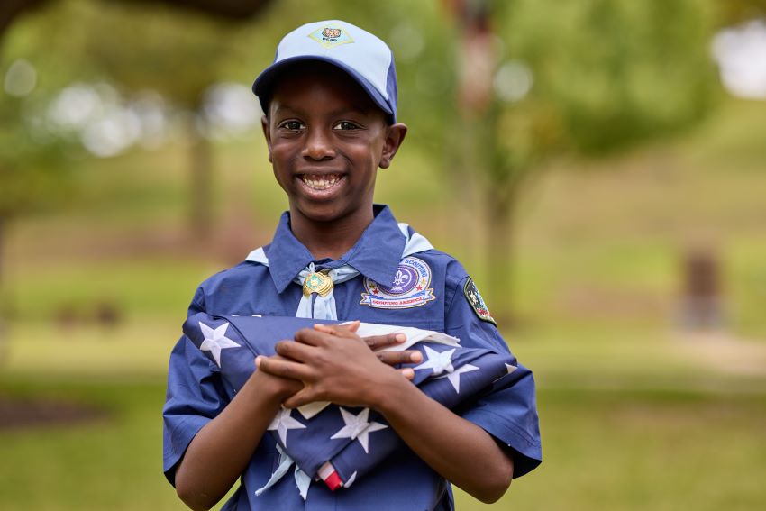 A black child in Cub Scout uniform holding a folded American flag outdoors